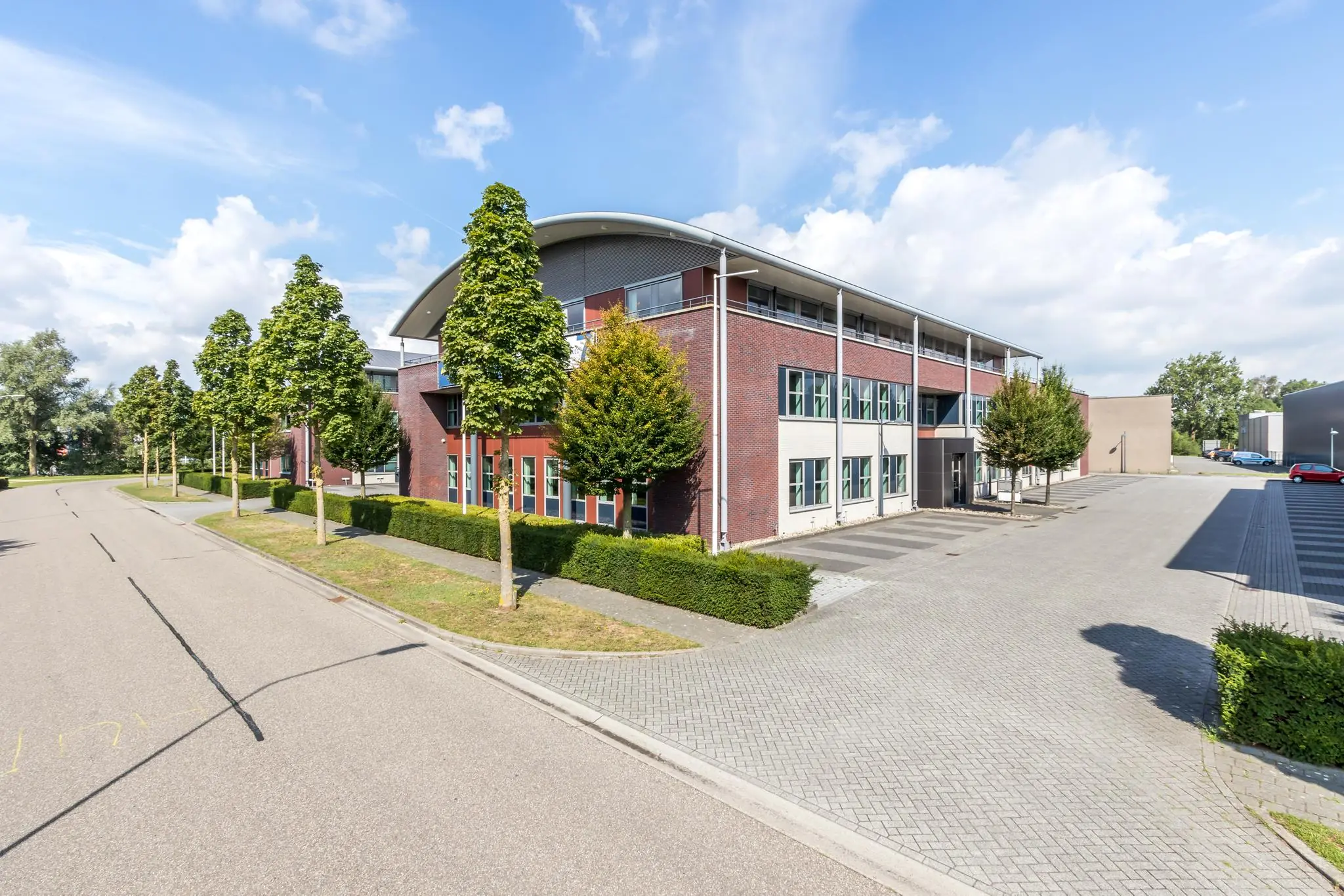 Modern kantoorgebouw aan de De Wel met rode en grijze gevel, omgeven door bomen en parkeerplaatsen.